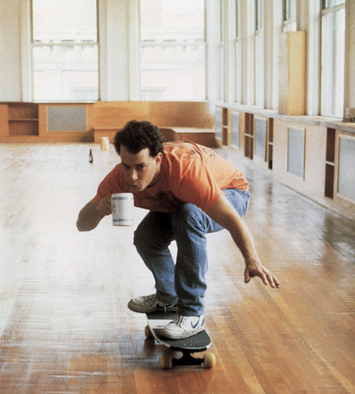 A man in an orange shirt mid-fall on a skateboard, coffee mug in hand, across an empty loft floor.